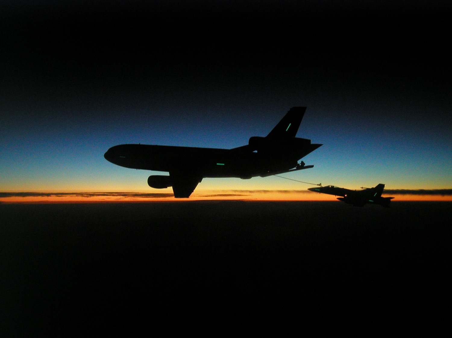A KC-10 tanker and an F/A-18 Hornet silhouetted against a sunset, the Hornet plugged into the refueling drogue.
