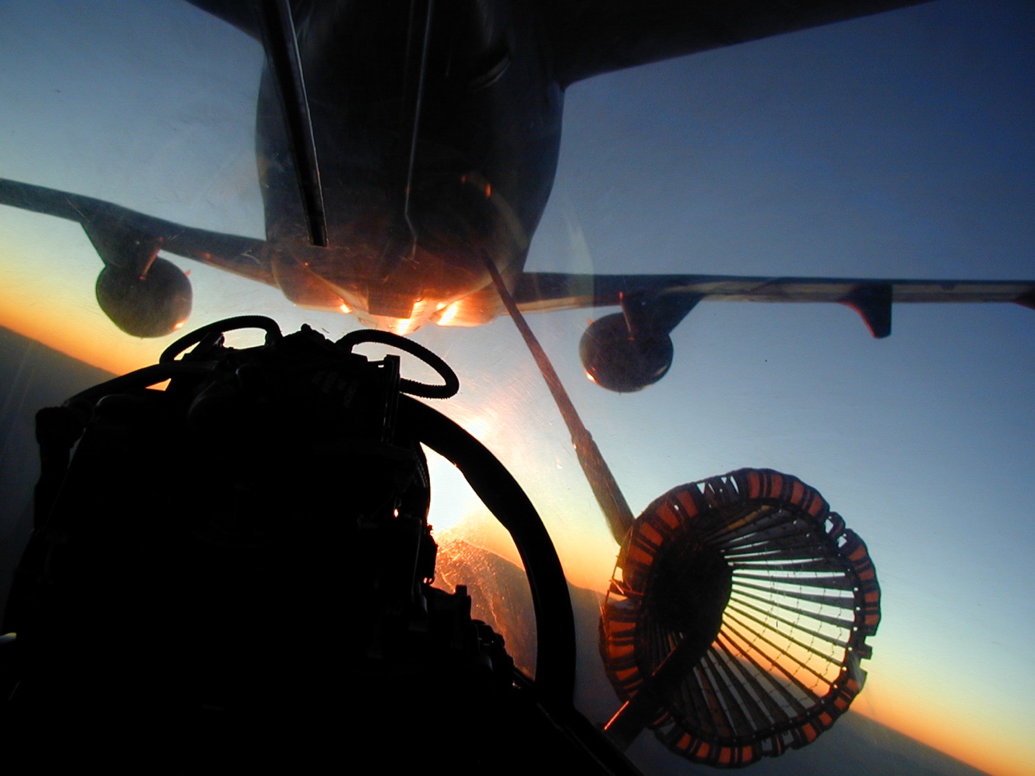 Cockpit view looking up at the tanker's belly with the refueling basket trailing on its hose, sun low on the horizon.