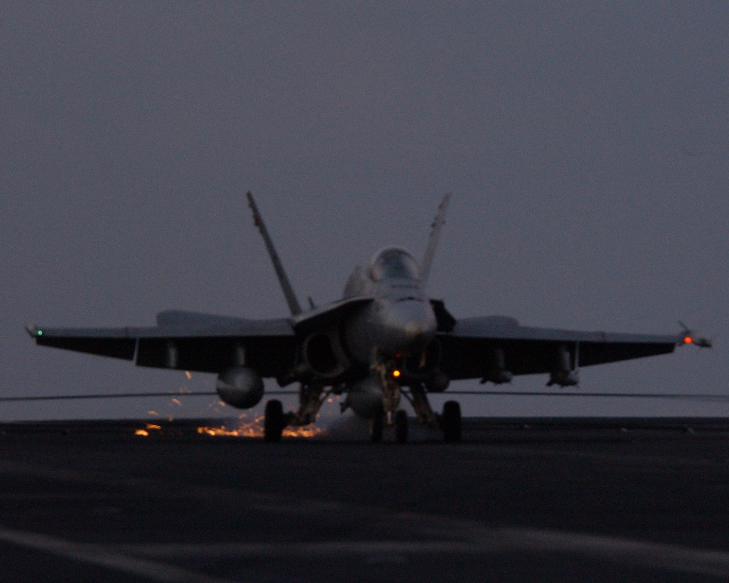 An F/A-18 Hornet catching the arresting wire at dusk, sparks streaming off the deck.
