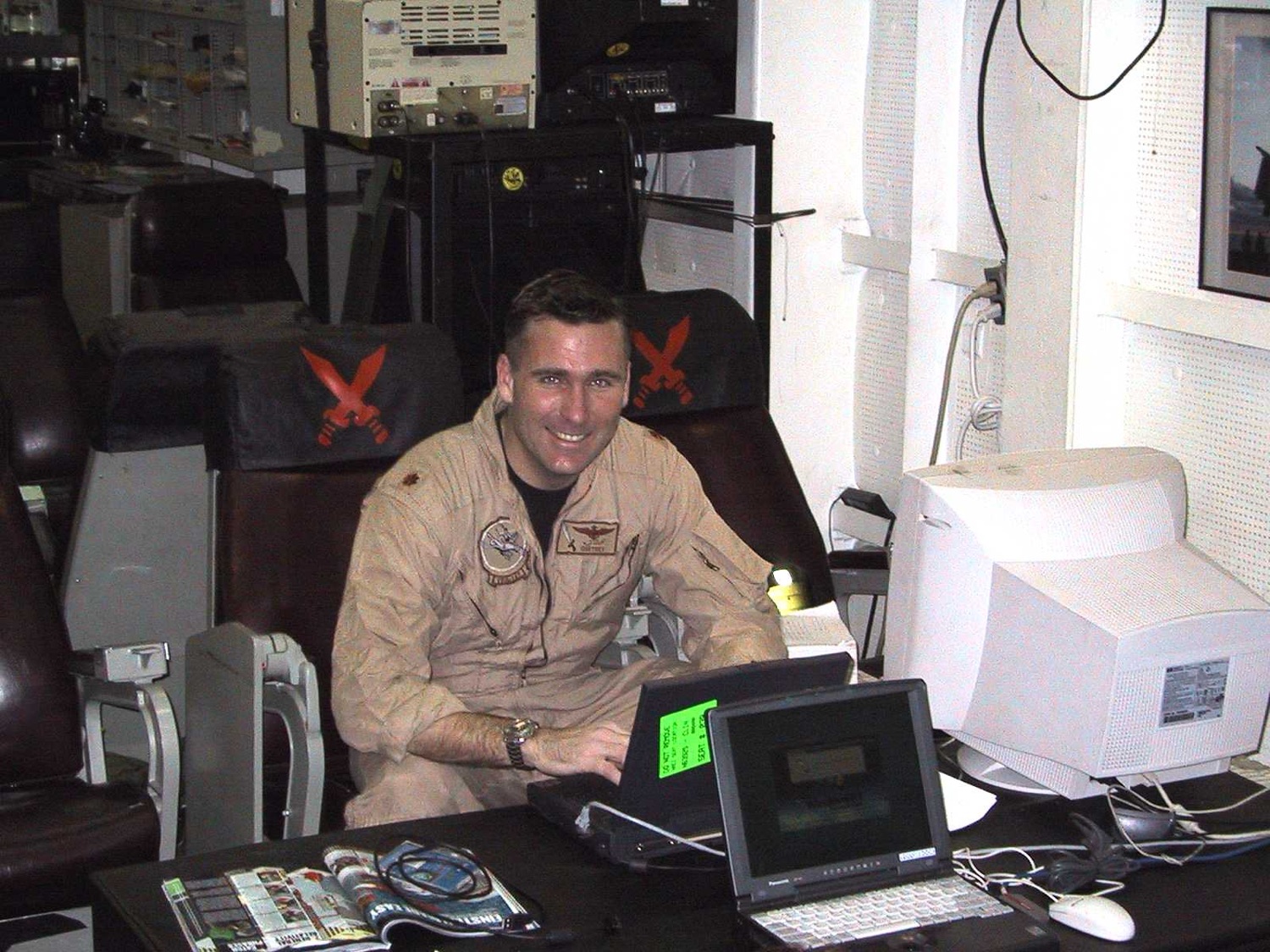 The author at the ready-room duty desk in a tan flight suit, laptop open, grinning, squadron chairs behind him.