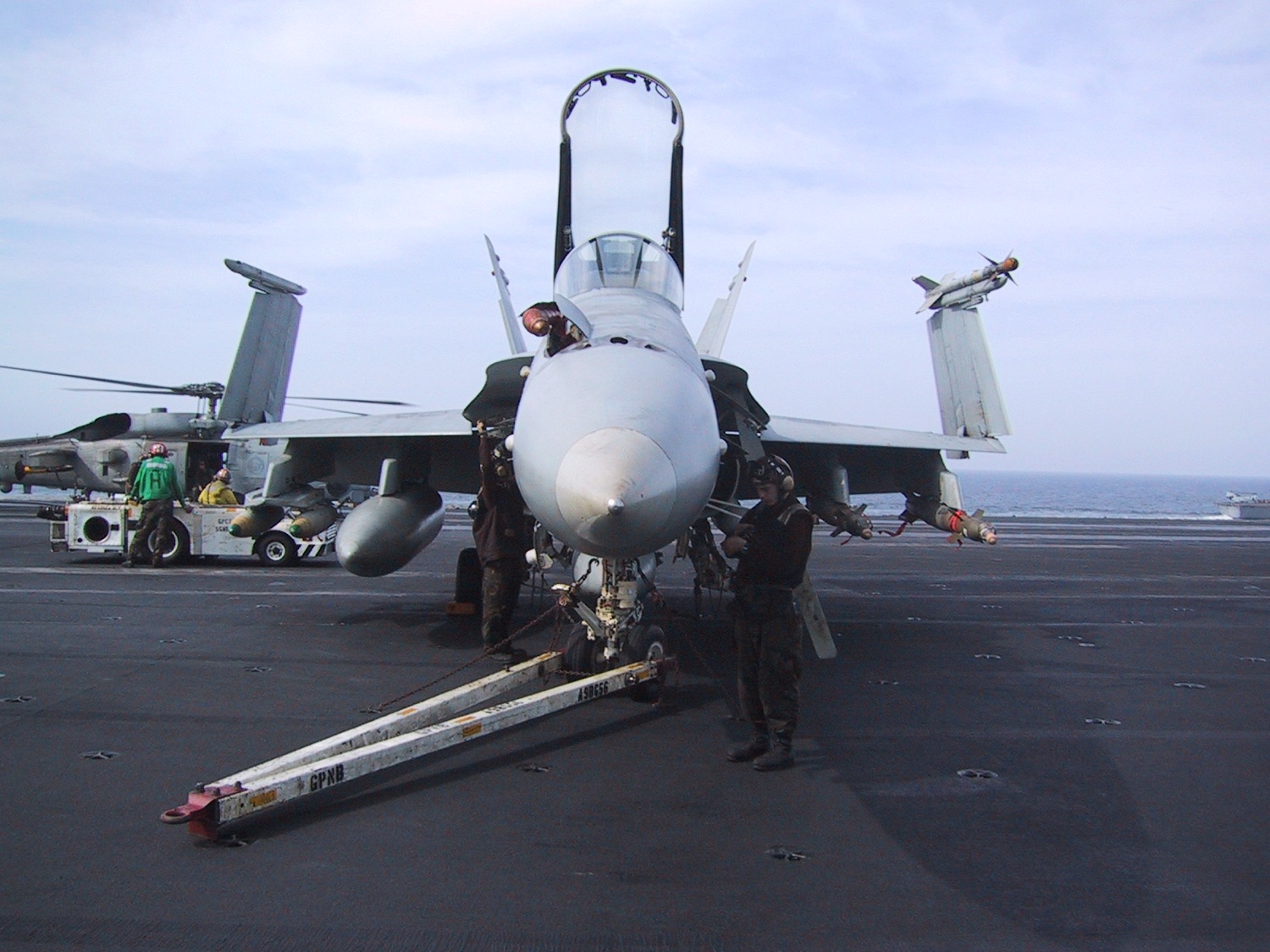 An F/A-18 Hornet on the carrier deck, launch bar down and crew at the nose, a helicopter parked behind.