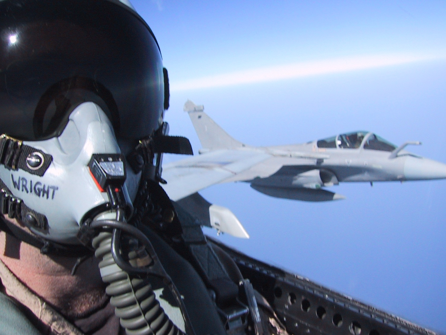 An F/A-18 launching off the bow low over the water while another Hornet sits parked, a warship in the distance.