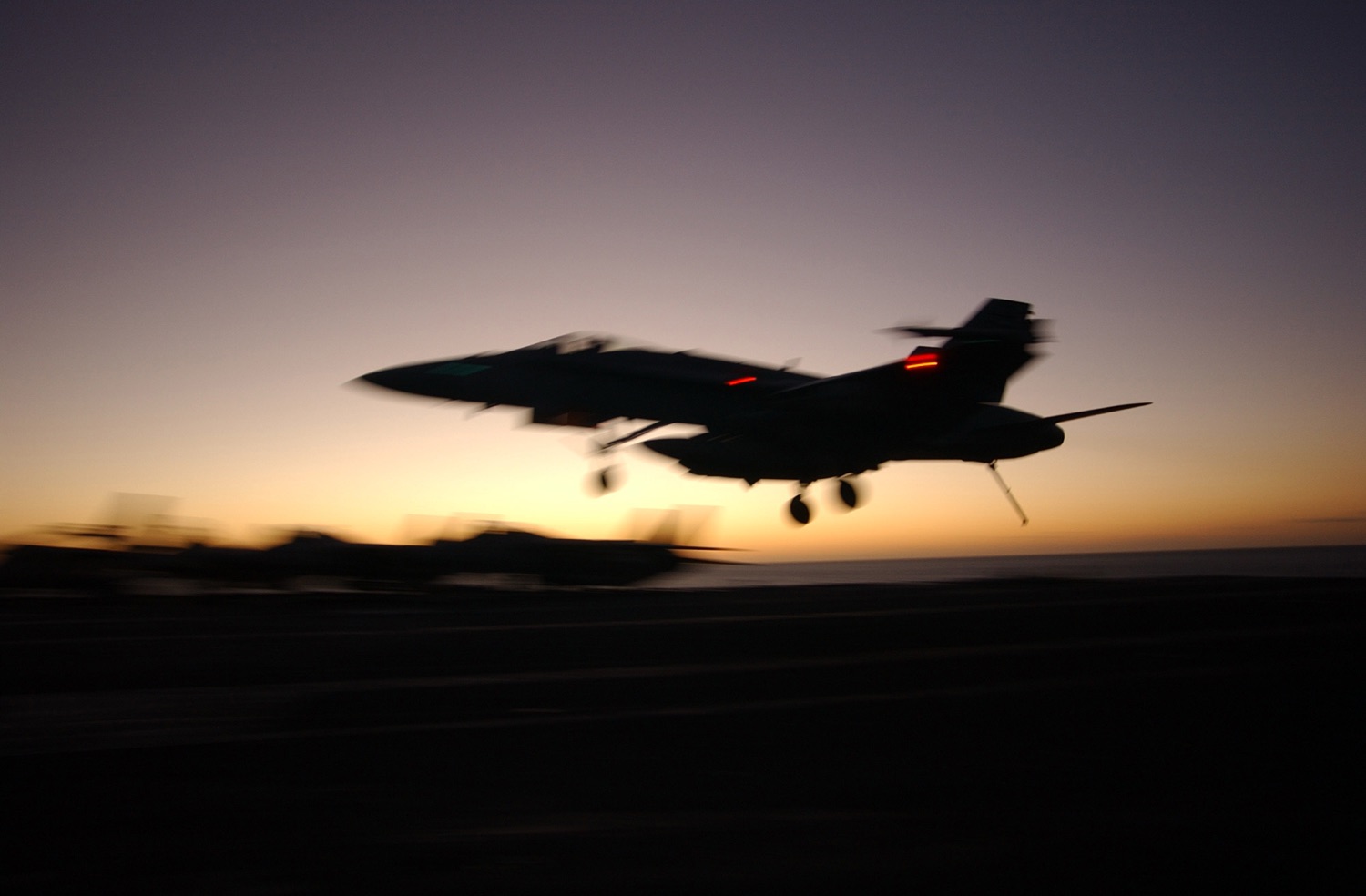 An F/A-18 Hornet on approach at dusk, motion-blurred against the last light on the horizon.