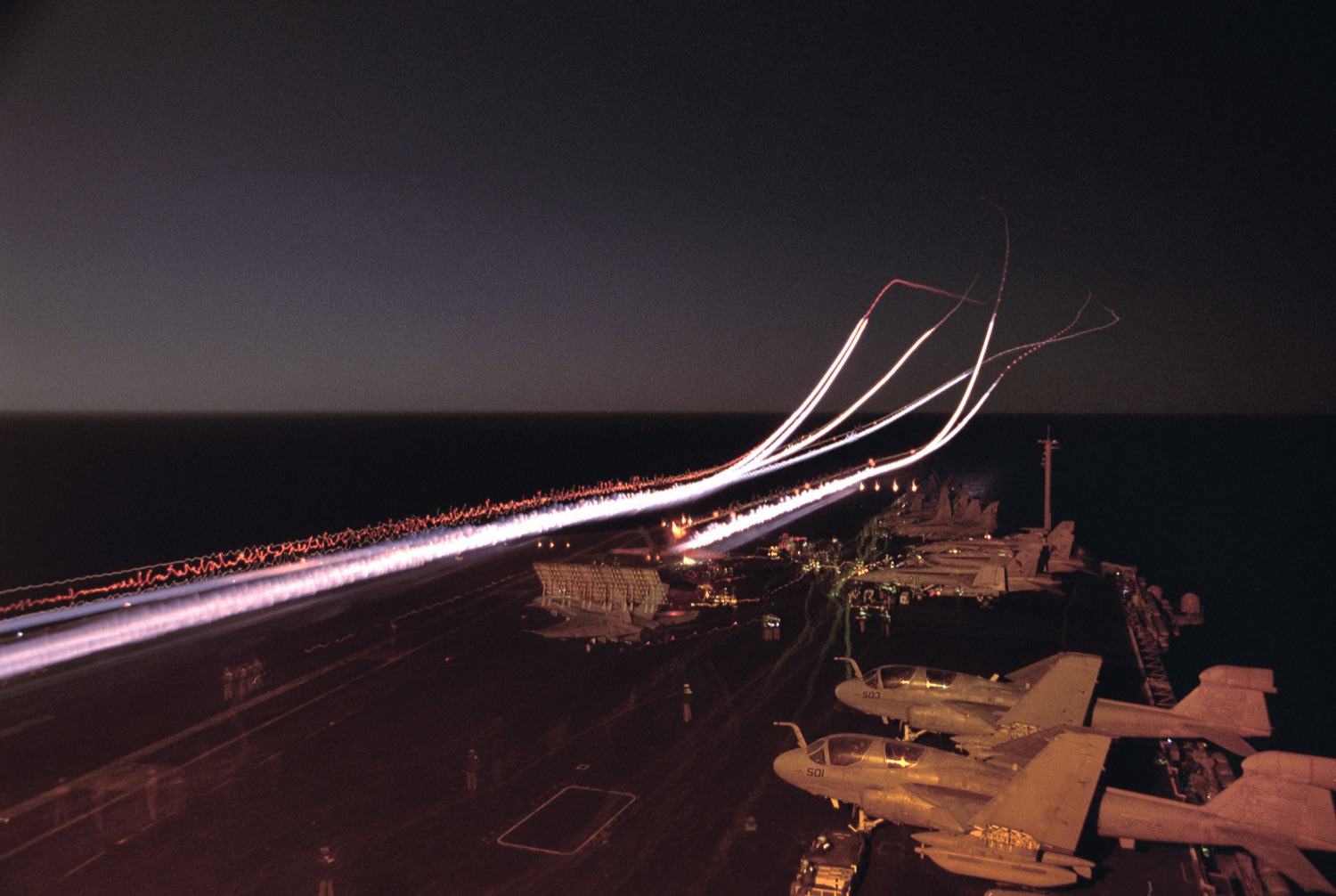 Night long-exposure of the carrier deck — light streaks from aircraft launching and recovering, Prowlers parked in the foreground.